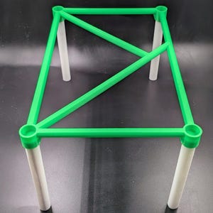 May include: A green geometric structure with a rectangular top and four white cylindrical legs. The top frame is a bright green color, with diagonal supports. The legs are white and evenly spaced, creating a stable base.