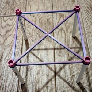 May include: A geometric structure with a square frame and four white legs. The frame is constructed with pink and blue rods, connected at the corners with pink cylindrical pieces. The structure is set on a wooden surface.