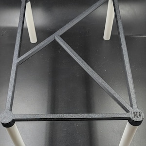 Pode incluir: Um suporte geométrico cinza escuro com uma estrutura retangular e pernas cilíndricas brancas. A estrutura tem um design de treliça e é suportada por quatro pernas. O suporte foi projetado para segurar um objeto.