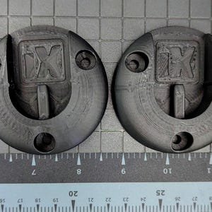 May include: Two dark grey, circular objects with a recessed centre and a handle-like projection. Each has the letters "IX" embossed in the centre and several holes around the perimeter. A ruler with inch and cm markings is in the foreground.
