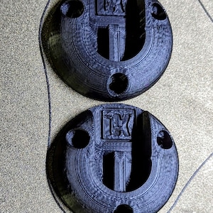 以下が含まれることがあります： 2つの黒い円形の3Dプリントコンポーネント。それぞれに中央のくぼんだ領域、3つの小さな穴、文字「TK」の隆起した長方形部分があります。コンポーネントはテクスチャ加工された表面にあります。