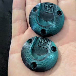 May include: Two teal-colored, circular objects with a textured surface and a raised design. Each has three small holes and a recessed square with the letters "IX". The objects are held in a hand.