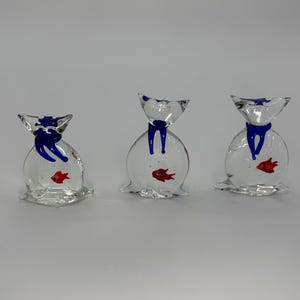 Puede incluir: Tres figuritas de vidrio con forma de bolsa con lazos azules y un pez rojo dentro de cada bolsa.