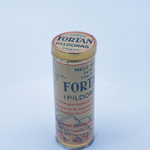 Può includere: Una latta cilindrica vintage con coperchio e base dorati, con la scritta "FORTAN PILDORAS" in un design circolare. Il corpo della latta è beige con testo stampato in spagnolo, tra cui "FORTAN (PILDORAS)" e altri dettagli.