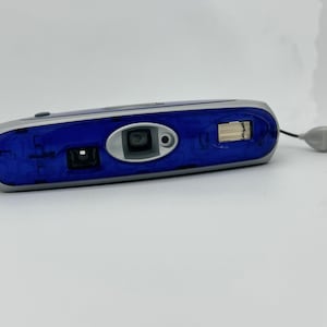 Puede incluir: Una cámara compacta azul y plateada con una tapa de lente y una correa de muñeca.