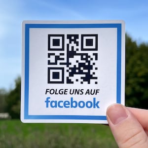 Könnte beinhalten: Ein weißer quadratischer Aufkleber mit einem schwarzen QR-Code und einem blauen Rand. Der Text "FOLGE UNS AUF facebook" ist in Schwarz unter dem QR-Code gedruckt.