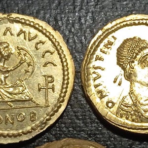Puede incluir: Dos monedas de oro con grabados detallados. Una moneda presenta una figura sentada con un escudo y la inscripción "VICTORIA AVGGG". La otra moneda muestra un perfil y la inscripción "DNIVSTINI ANVSPPAVG".