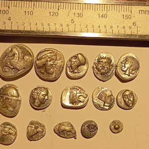 Puede incluir: Una colección de 14 monedas plateadas con varios diseños, incluyendo un león, un caballo, una cara humana y un pájaro. Las monedas están dispuestas en tres filas sobre una superficie marrón claro.