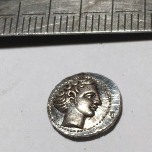 Puede incluir: Moneda de plata con un perfil de una persona con el pelo rizado en un lado. La moneda mide aproximadamente 2,5 cm de diámetro.