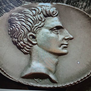 May include: A silver-toned metal medallion with a profile portrait of a man with short, wavy hair. The medallion has a beaded edge.
