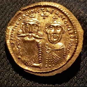 Op de afbeelding: Een gouden munt met twee gekroonde figuren met gedetailleerde gelaatstrekken en inscripties. De munt vertoont tekenen van slijtage en ouderdom. Het ontwerp van de munt omvat ingewikkelde details en een getextureerd oppervlak.