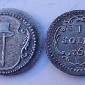 Peut inclure: Une pièce d'argent avec un dessin d'un chapeau sur un bâton d'un côté et le texte "J SOLD0 J769" de l'autre côté.