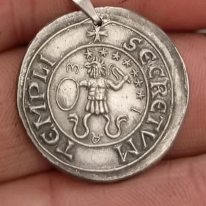 Pode incluir: Um pingente de prata com um design circular que apresenta uma figura segurando uma espada e um escudo. O pingente tem a inscri&ccedil;&atilde;o "SECRETVM LEGATI" ao redor da borda.