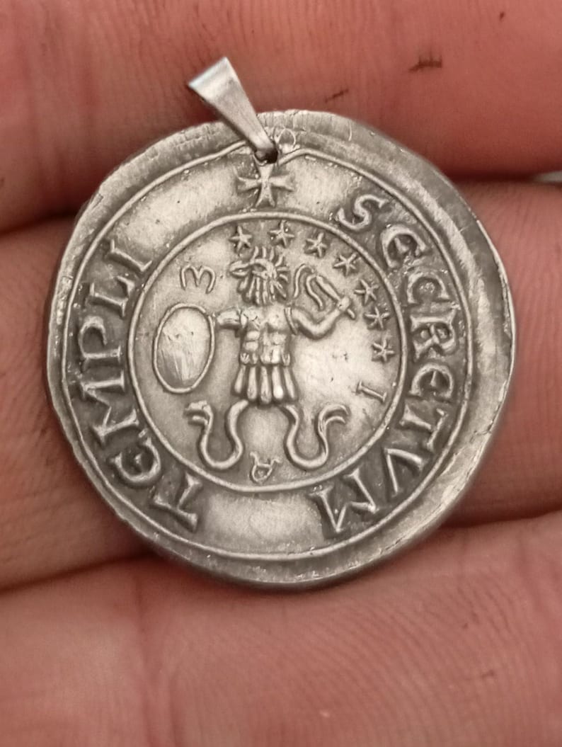 Pode incluir: Um pingente de prata com um design circular que apresenta uma figura segurando uma espada e um escudo. O pingente tem a inscri&ccedil;&atilde;o "SECRETVM LEGATI" ao redor da borda.
