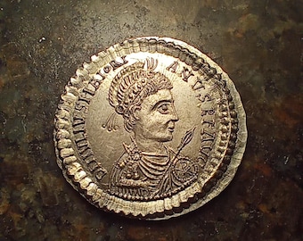 May include: A silver coin featuring a profile of a person wearing a laurel wreath and armor, holding a spear and shield. The coin has detailed inscriptions around the edge and a textured border. The coin appears to be an ancient artifact.