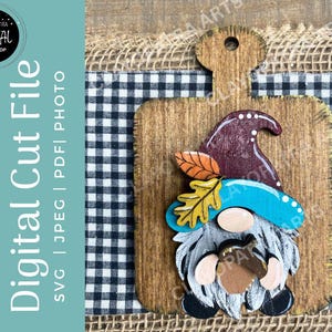 May include: A wooden gnome decoration with a blue hat, holding an acorn, and wearing a burgundy hat. The gnome is on a wooden cutting board with a black and white checkered background. The text "Digital Cut File" is on the left.