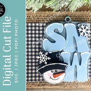May include: A wooden snowman-themed ornament with the word "SNOW" in light blue letters. The snowman has a black hat, a carrot nose, and a smiling face. A snowflake is placed above the "W". The background includes a blue and white checkered pattern.