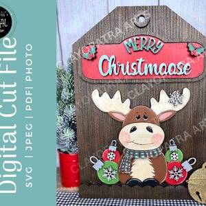 May include: Wooden Christmas decoration featuring a moose with antlers, a scarf, and ornaments. The sign reads "Merry Christmaase" in red and teal. The background is a wooden plank design with a small bell.