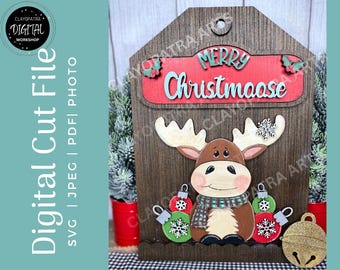 May include: Wooden Christmas decoration featuring a moose with antlers, a scarf, and ornaments. The sign reads "Merry Christmaase" in red and teal. The background is a wooden plank design with a small bell.