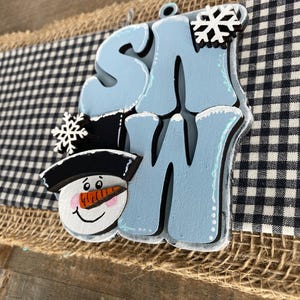 May include: A wooden sign with the word "SNOW" in light blue, featuring a snowman face and snowflakes. The sign is set against a backdrop of burlap and a black and white checkered pattern.