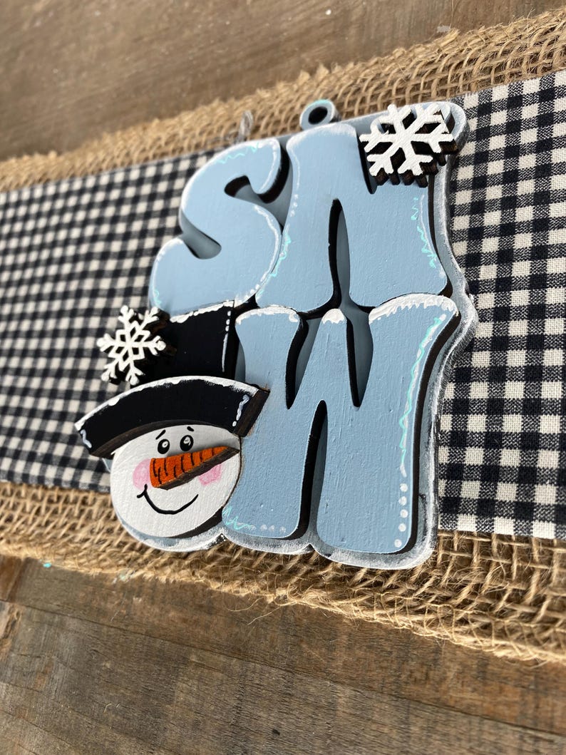 May include: A wooden sign with the word "SNOW" in light blue, featuring a snowman face and snowflakes. The sign is set against a backdrop of burlap and a black and white checkered pattern.