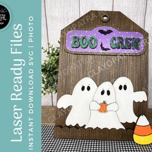 Könnte beinhalten: Eine Halloween-Dekoration aus Holz mit drei weißen Geisterfiguren und einem Zuckermais. Das Schild trägt die Aufschrift "BOO CREW" in grünen Glitzerbuchstaben auf lila Hintergrund. Der Hintergrund hat ein Holzmaserungsmuster.