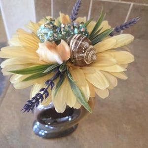 May include: A decorative floral arrangement featuring a large yellow daisy as the base. Adorned with a seashell, lavender sprigs, and faux greenery. The arrangement is set in a brown ceramic vase, creating a coastal or beach-themed aesthetic.