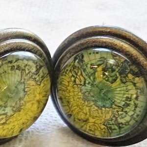 May include: A collection of decorative cabinet knobs. Some are light green ceramic with bronze accents, while others feature a map design under glass with bronze-colored metal. A ruler is placed below the knobs.