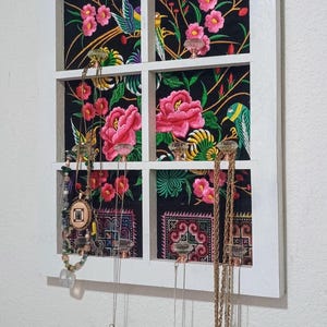 May include: White framed jewelry organizer with a floral fabric backing. The fabric features pink flowers, green leaves, and birds on a black background. Various necklaces hang from the frame, showcasing a variety of pendants.