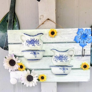 May include: A white wooden sign with two small blue and white porcelain pitchers and bowls. The sign is decorated with artificial sunflowers, daisies, and a blue flower with a hook. Decorative metal musical instruments hang on either side.
