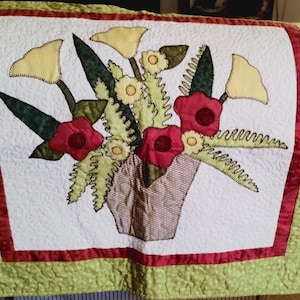 May include: A white quilted wall hanging with a green and red border. The center features a brown pot with a bouquet of yellow and red flowers.