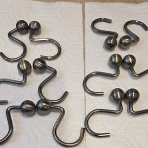 May include: A collection of shower curtain hooks. Each hook is made of metal and features a decorative ball at the top. The hooks are arranged on a white surface.