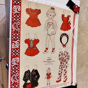 May include: A framed vintage paper doll print featuring a young girl and various outfits in red and black. The print is decorated with red paisley ribbon, white lace, and a black fringe.