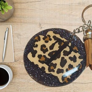 Könnte beinhalten: Ein runder, schwarzer Glitzer-Schlüsselanhänger mit einem braunen und schwarzen Leopardenmuster. Der Schlüsselanhänger hat einen silbernen Metallring und einen braunen Quasten.