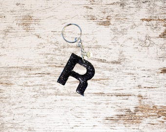 May include: Black glitter resin letter "R" keychain with a silver keyring and chain. The letter is attached to a small charm. The background is a white and brown distressed wood.