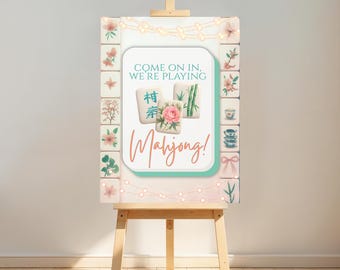 Könnte beinhalten: Eine hölzerne Staffelei zeigt ein Schild mit den Worten "Come on in, we're playing Mahjong!" in einer dekorativen Schriftart. Das Schild zeigt Blumen- und Mahjong-Kachel-Illustrationen mit einer Lichterkette oben.