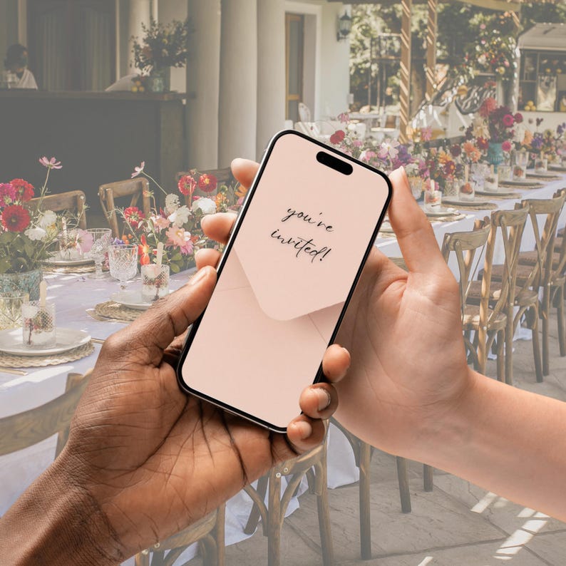 May include: A person holding a smartphone with a pink screen that says "you're invited!" The phone is in front of a blurred background of a table setting with flowers and chairs.