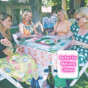 K&ouml;nnte beinhalten: Eine Gruppe von vier Personen spielt Mahjong an einem Tisch mit einer gebl&uuml;mten Tischdecke. Das Spielset besteht aus gr&uuml;nen und wei&szlig;en Spielsteinen auf einem Brett. Ein Schild mit dem Text Perfect for Mahjong Classes ist im Vordergrund zu sehen.
