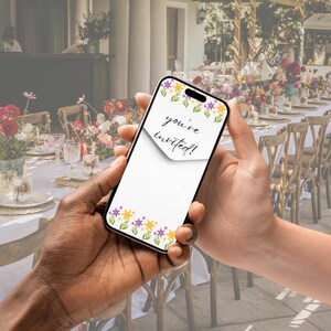 May include: A person holding a smartphone with a floral design and the text "you're invited!" on the screen. The phone is being held in front of a table with white linens and floral centerpieces.