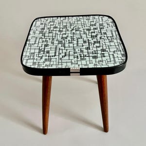 Könnte beinhalten: Ein kleiner quadratischer Beistelltisch mit einem schwarz-weißen geometrischen Muster auf der Oberseite. Der Tisch hat einen schwarzen Rand und drei Holzbeine.