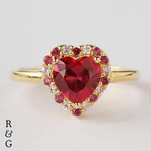 Op de afbeelding: Een gouden ring met een grote rode hartvormige edelsteen, omringd door kleinere roze en witte edelstenen.