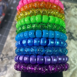 May include: A stack of colorful beaded bracelets in a rainbow pattern. The bracelets are made of small, round beads with a glittery finish. The beads are arranged in a stack, with the pink bracelet on top and the purple bracelet on the bottom.