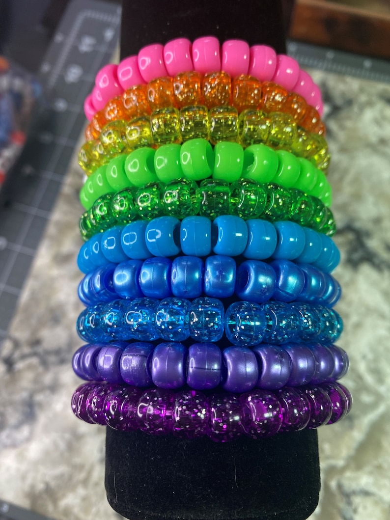 May include: A stack of colorful beaded bracelets in a rainbow pattern. The bracelets are made of small, round beads with a glittery finish. The beads are arranged in a stack, with the pink bracelet on top and the purple bracelet on the bottom.