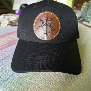 May include: Black baseball cap with a curved brim. The cap features a circular, two-tone brown and tan leather patch with a stylized design. The cap is made of a textured fabric.
