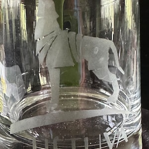 May include: Clear glass tumbler with etched design. The design features a stylized figure and the word "LITHUANIA" etched at the base. The glass has a cylindrical shape and is suitable for beverages.
