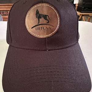 以下が含まれることがあります： カーブしたつばが付いた濃い茶色の野球帽。オオカミのシルエットと白い文字で「LIETUVA」と書かれた円形の革製パッチが付いています。パッチは帽子に縫い付けられています。
