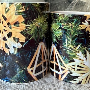 May include: Two white ceramic mugs with a Christmas tree design. The mugs feature a close-up of a Christmas tree with straw ornaments, including snowflakes and stars.