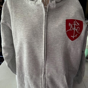Può includere: Una felpa con cappuccio grigio chiaro con cerniera e tasche anteriori. La felpa ha un emblema rosso sul petto sinistro. L'emblema ha la forma di uno scudo con un disegno di spade e altri simboli.