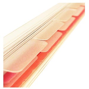 May include: Close-up of a clear, plastic binder with several rectangular, translucent tabs. The tabs are a light beige color and are layered over a red and white background. The binder is angled diagonally.