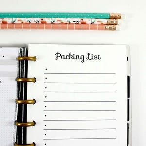 May include: A planner with a "Packing List" page, gold binder rings, and decorative pencils. Pink and teal binder clips and a floral magnet are also present. The items are arranged on a white surface, creating a clean and organized aesthetic.
